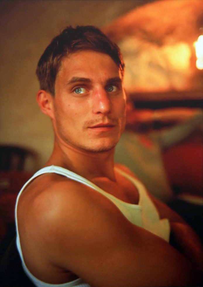 This is a color photographic portrait of a man with short blonde hair and a white tank top looking to his right past the camera.