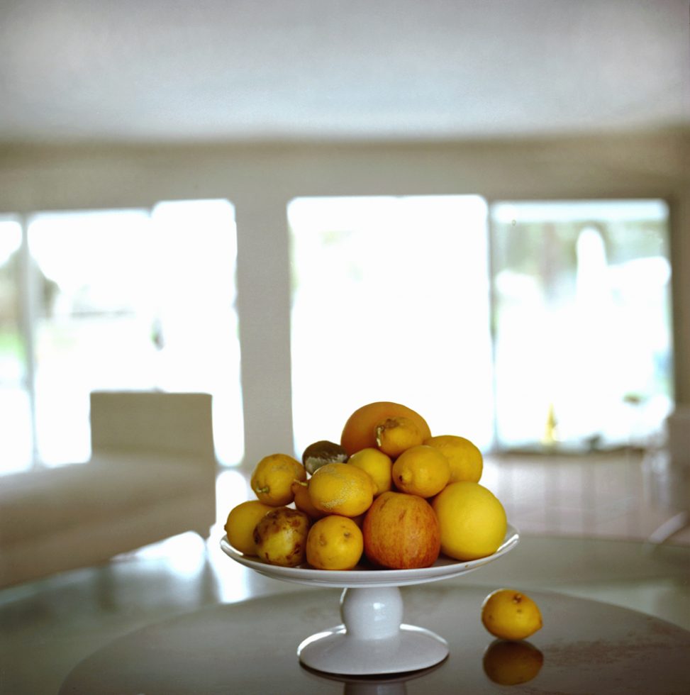 This is a color photograph of various orange and yellow fruits in a shallow bowl on a round table.