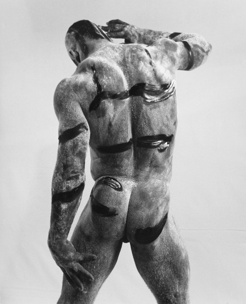 This is a black-and-white photograph of the backside of a naked Black man with paint on his skin.