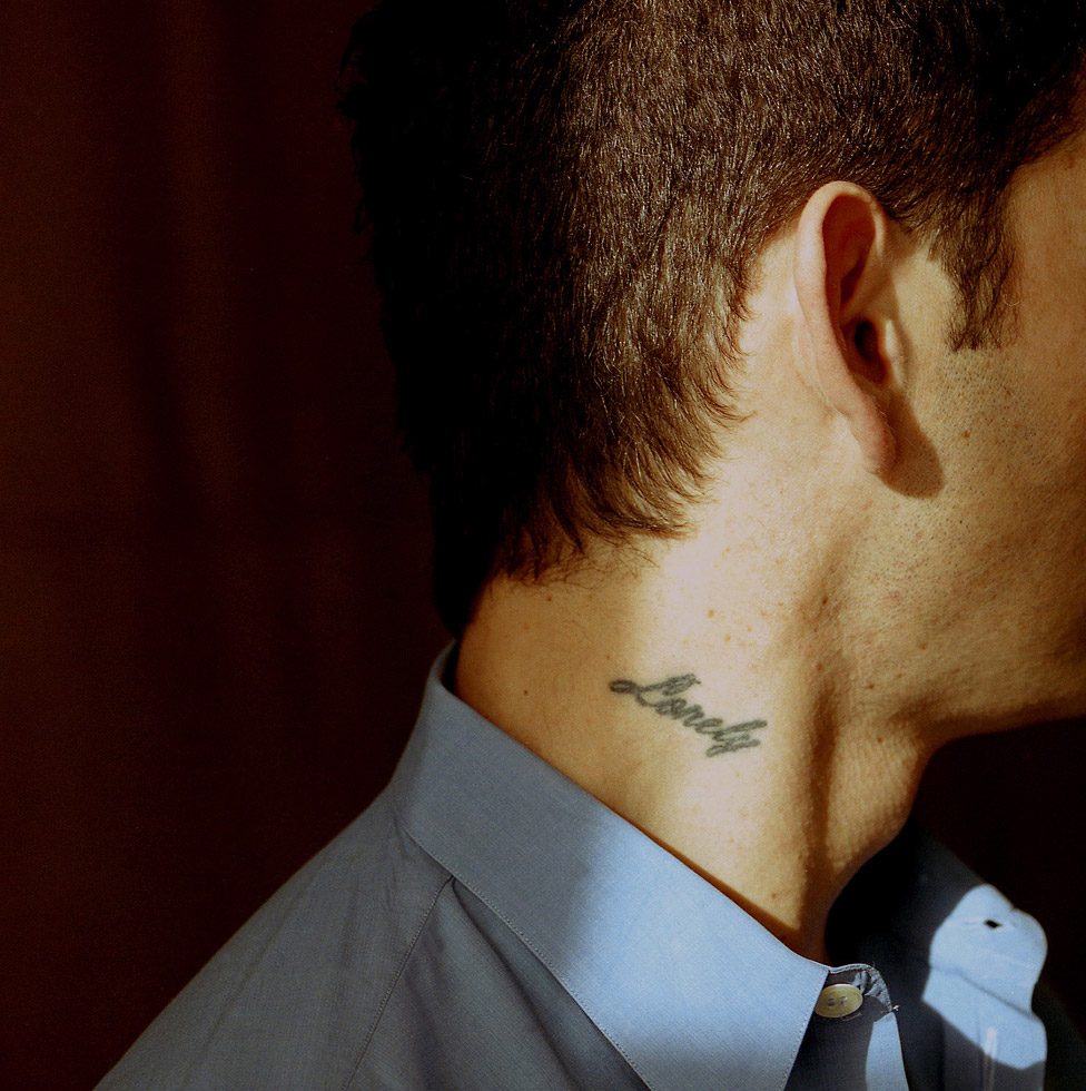 This is a color photograph of a man in a blue collared shirt in profile with a neck tattoo that reads "lonely."