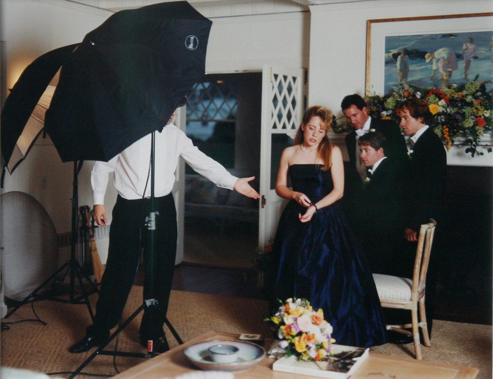 This is a color photograph of a woman in a formal blue dress with three men in tuxedos and a wedding photographer whose head is obscured by a flash umbrella.