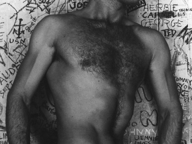 This is a black-and-white photograph of a shirtless man against a graffiti background pulling down his white pants.