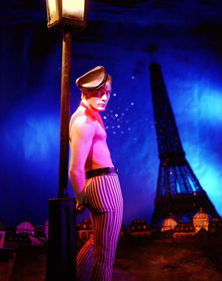 James Bidgood photograph of Apache played by model Tommy Coombs standing by a lamppost in pink light in front of the Eiffel tower and blue sky.