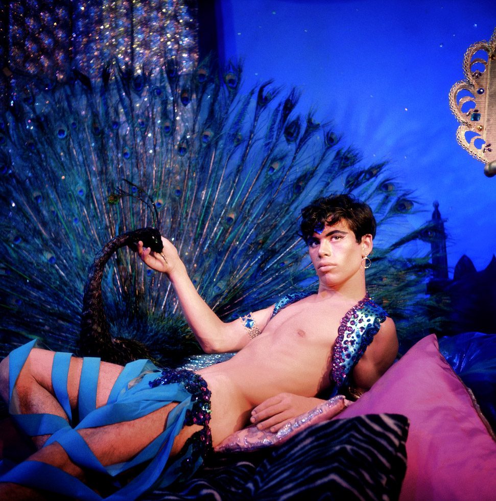 A photograph by James Bidgood of a shirtless harem boy in font of a peacock in a blue room