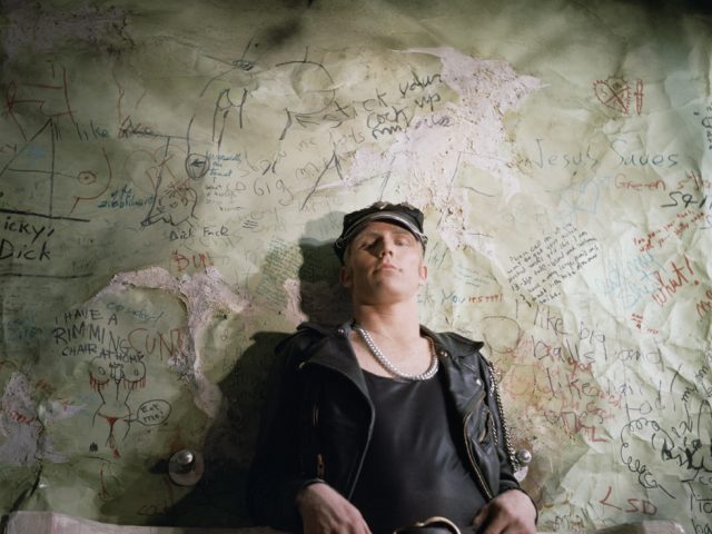 A photograph by James Bidgood of a man in leather jacket leaning against urinals with graffiti