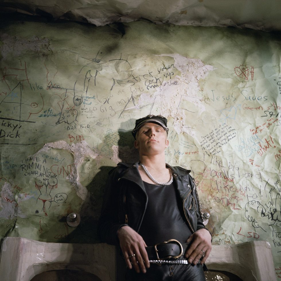 A photograph by James Bidgood of a man in leather jacket leaning against urinals with graffiti