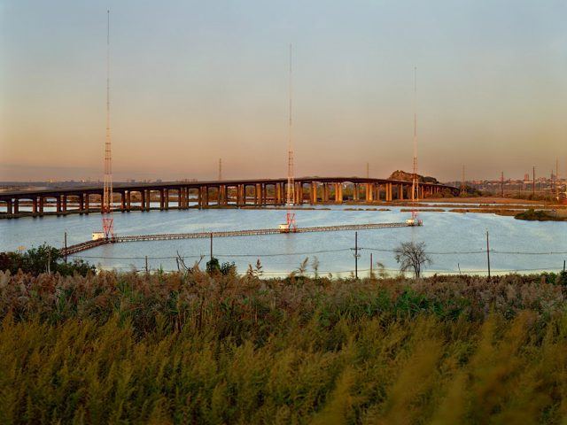 This is a color photograph of a blue body of water between a green hill and a bridge.