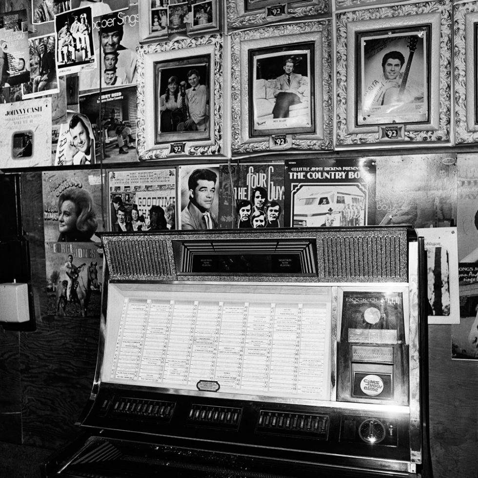 This is a black-and-white photograph of a jukebox with old photographs hanging above it.