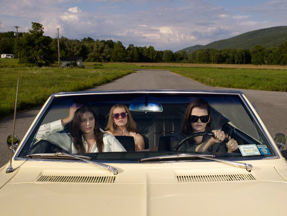 This is a color photograph of three young women seen through the windshield of a yellow convertible car.