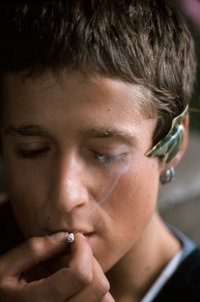 A close up photograph of a young person smoking, with a leaf tucked behind their ear