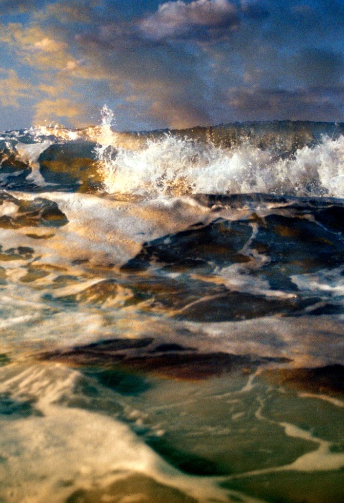 A photograph of a wave crashing with the glow of a sun set reflecting in the water