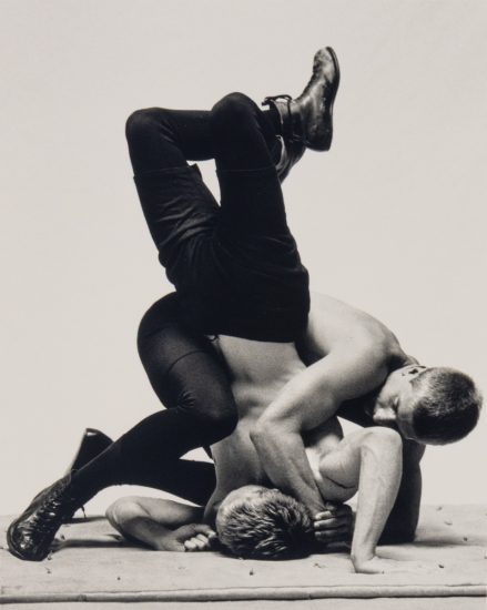 This photograph shows two men wrestling with one another. One pins the other against the ground.