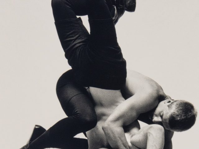 This photograph shows two men wrestling with one another. One pins the other against the ground.