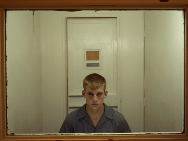 A color photograph of a blond young man in a prison behind a framed sheet of glass.
