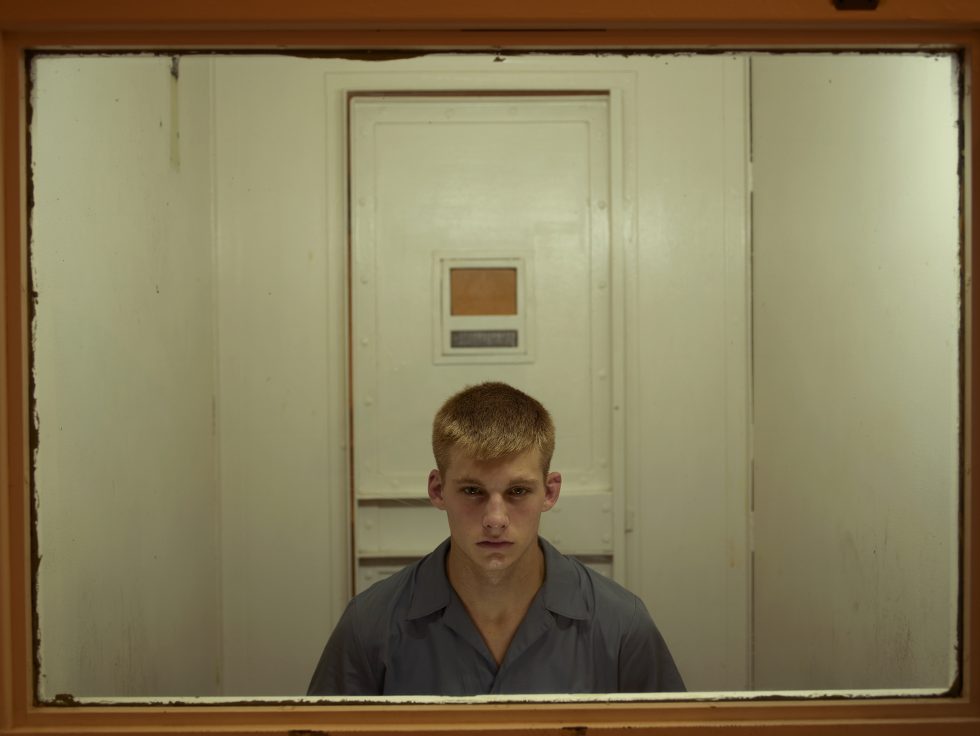 A color photograph of a blond young man in a prison behind a framed sheet of glass.