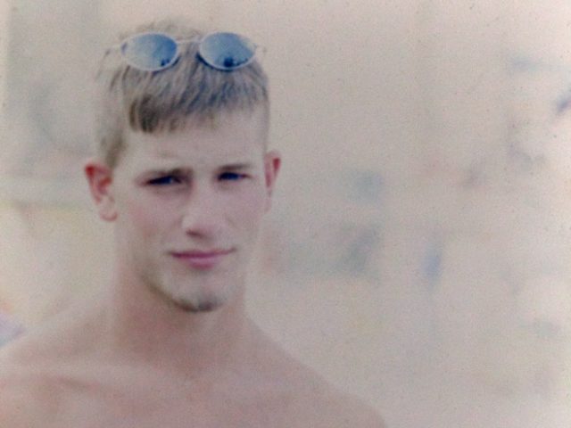 Photograph of a young man with blonde hair and a goatee with sunglasses resting on top of his head