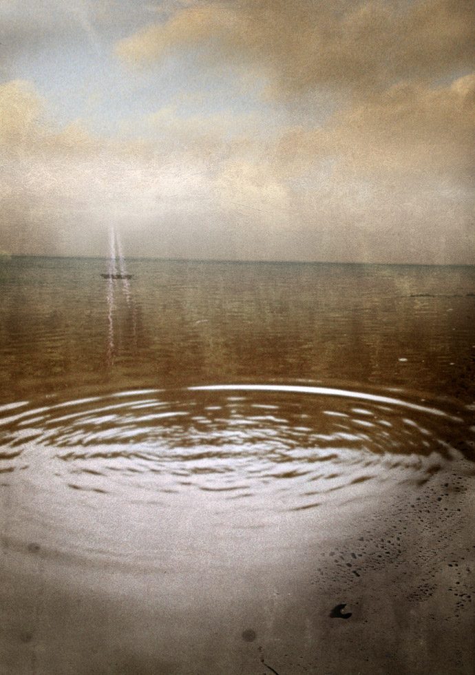 A hazy photograph of a sailboat out in the distance of the ocean, in front of the camera is a circular ripple in the water