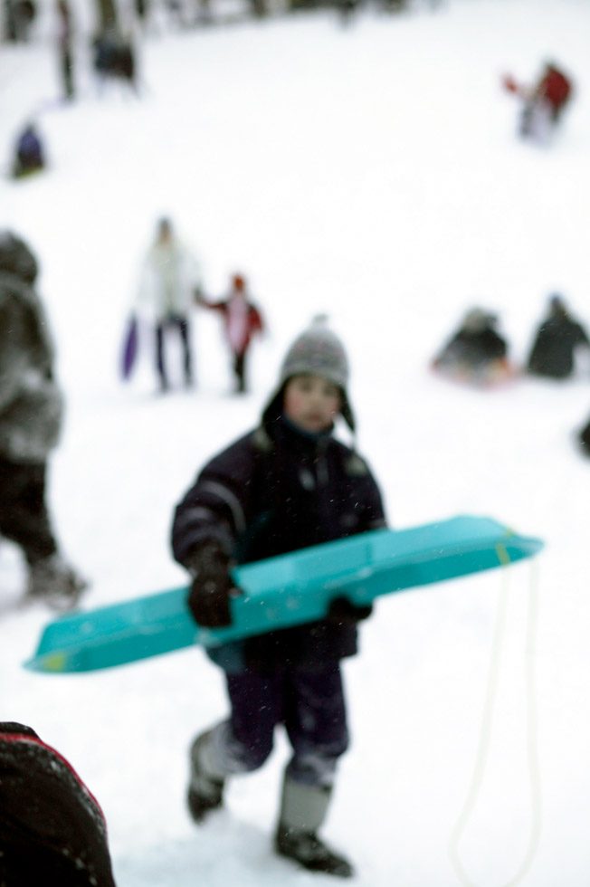 A soft focus photograph of a child climbing up a snowy hill carrying a teal sled