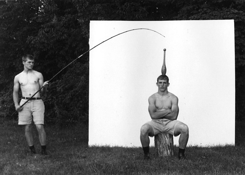 This is a black-and-white photograph of two young men, a fishing rod, and an Indian club.