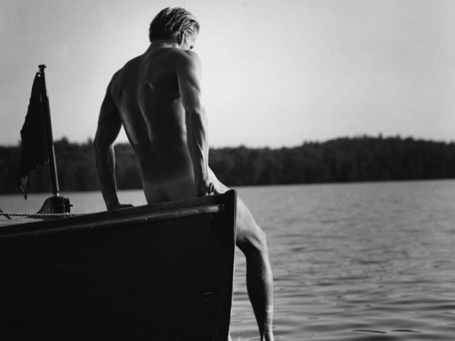 Photograph of a man sitting with his back turned to the camera on the edge of a boat with a foot dangling in the water