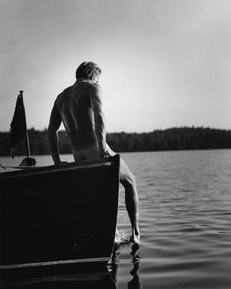 Photograph of a man sitting with his back turned to the camera on the edge of a boat with a foot dangling in the water