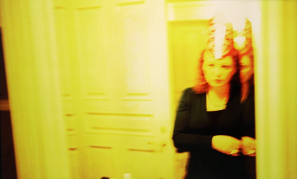 Photograph of a woman bathed in a yellow light looking at herself in a mirror wearing a tall party hat