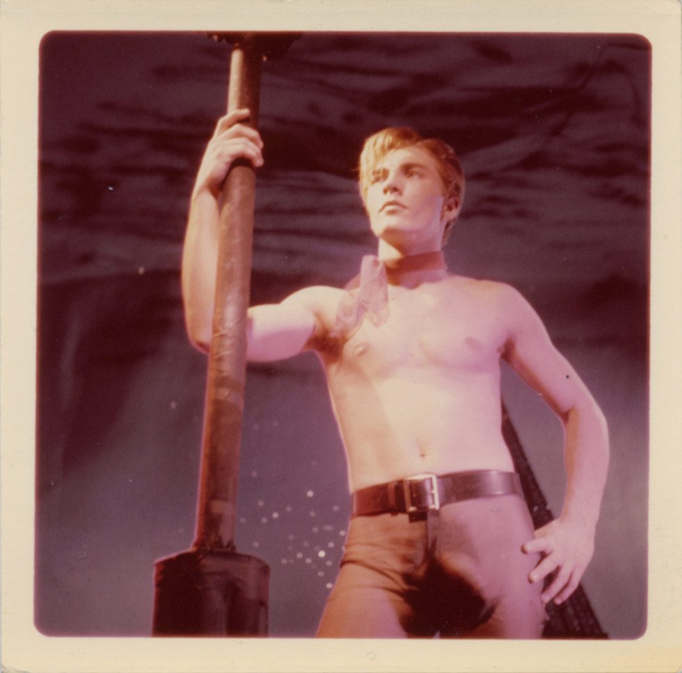 A James Bidgood photograph of a model named apache stanind shirtless with hand on hip leaning on a lamppost