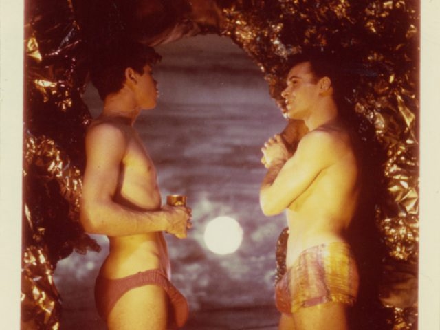 A James Bidgood photograph of two men in speedos standing by the opening of a cave with he moon shining