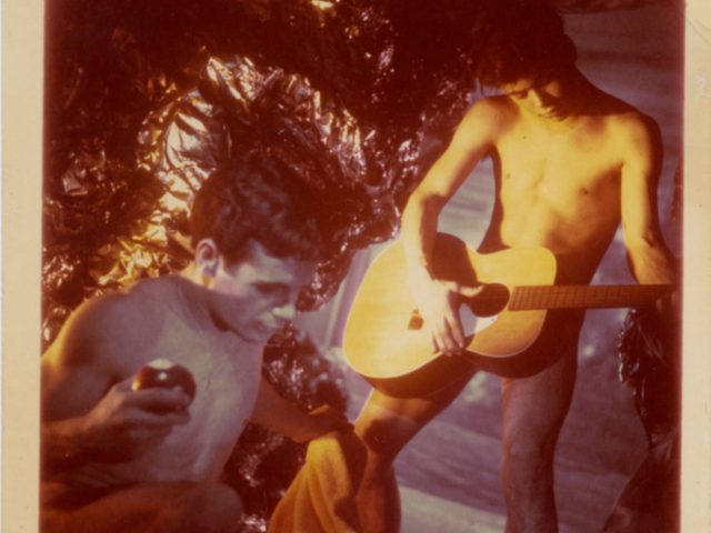 A photograph by James Bidgood of two boys in a cave. One is playing the guitar.