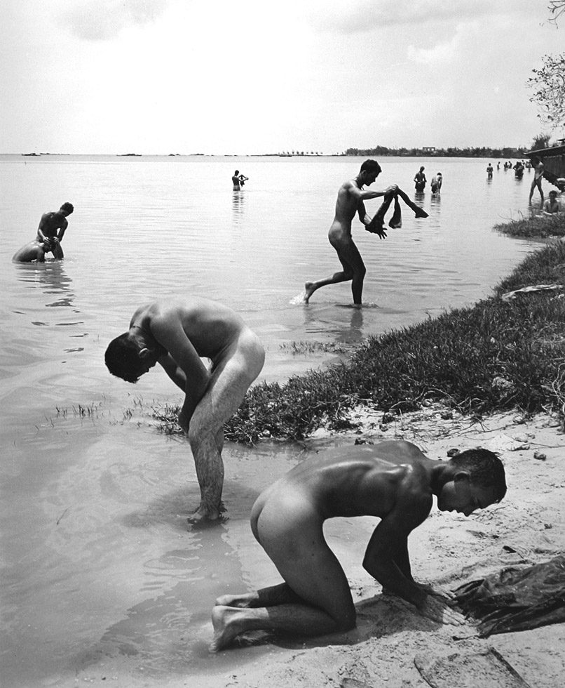 This is a black-and-white photograph of naked men on a sandy beach and in the water.