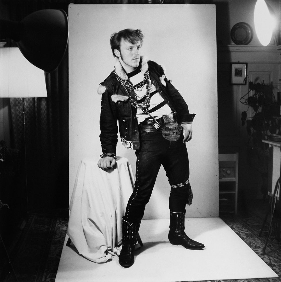 This is a black-and-white photograph of a teenage guy in a studio setting who is posing in jeans, boots, a jacket, and a big Elvis belt buckle.