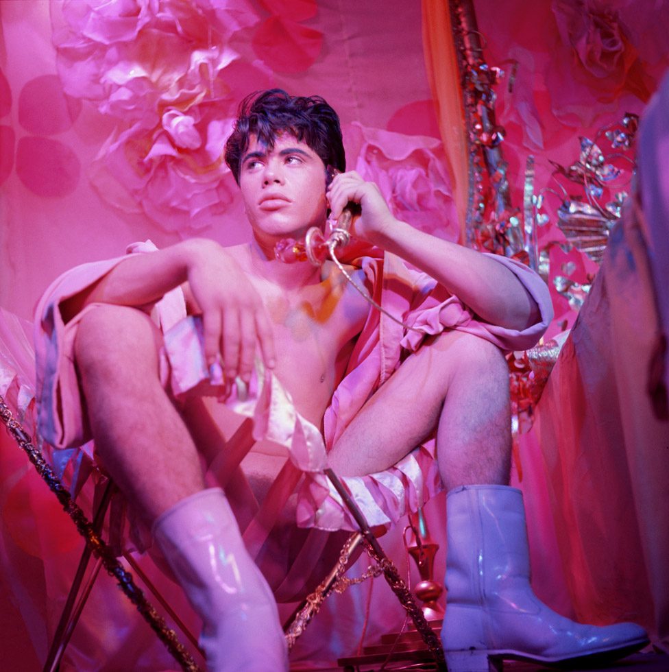 A photograph by James Bidgood of Bobby Kendall in white cowboy boots sitting in a chair holding a phone in a pink room