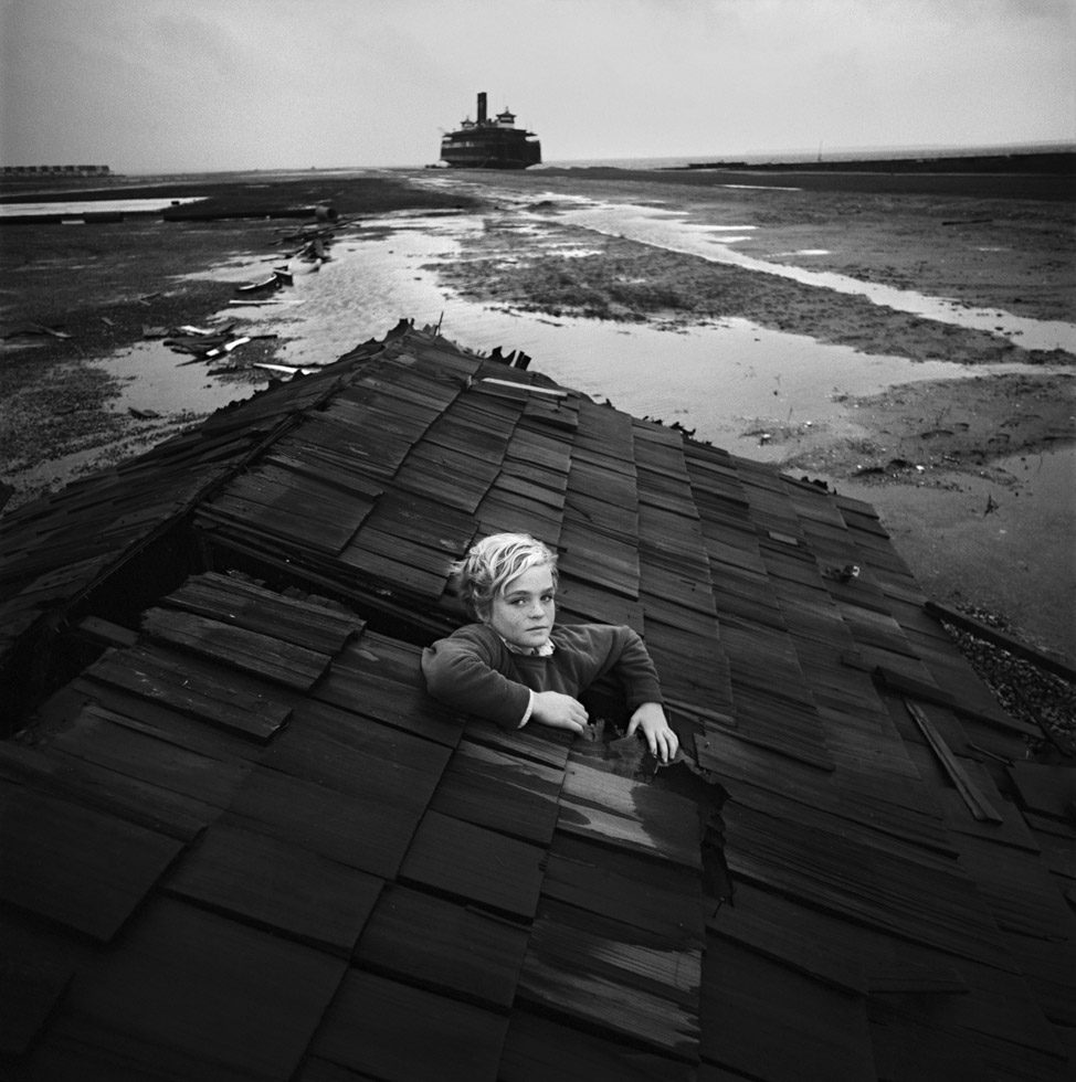 This is a black-and-white photograph of a young, blond child emerging from a hole in a roof on a beach with a ship on the horizon.