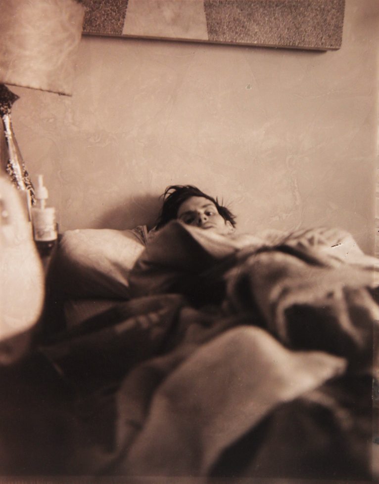 This is a sepia-toned photograph of a man in bed with the blankets pulled up to his chin.