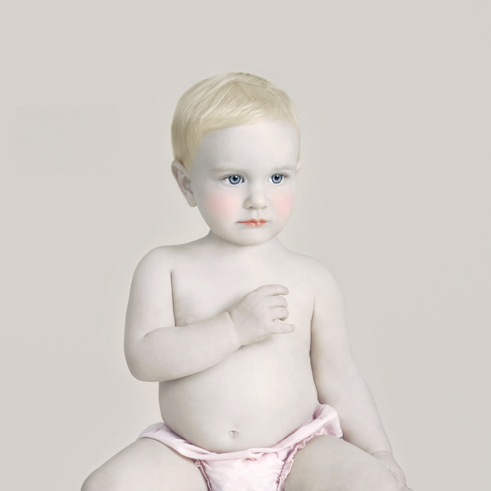 This is a color photograph of a baby in a pink diaper with her hand to her chest looking to the side.