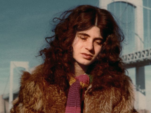 This is a color photograph of a young woman with thick, brown hair in a brown fur coat and knitted scarf in front of a white bridge.