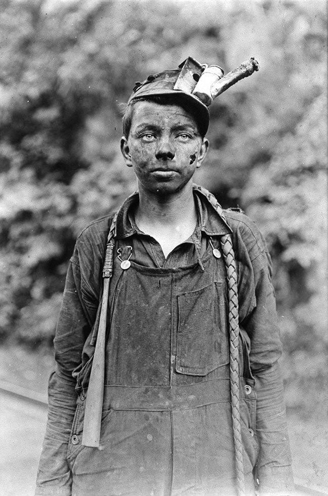 This is a black-and-white photograph of a young boy in overalls with his face covered in coal dust.