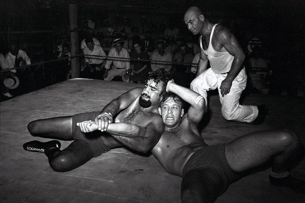 This is a black-and-white photograph of two wrestlers engaged in a ring with a referee looking on from the side.