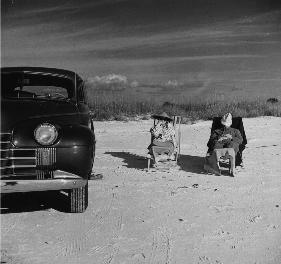 This is a black-and-white photograph of a man and a woman in deck chairs sitting in the sun in sand next to an old-fashioned car.