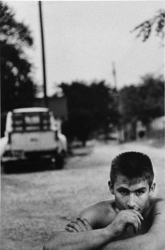 A black-and-white photograph of a shirtless young man on front of an old truck.