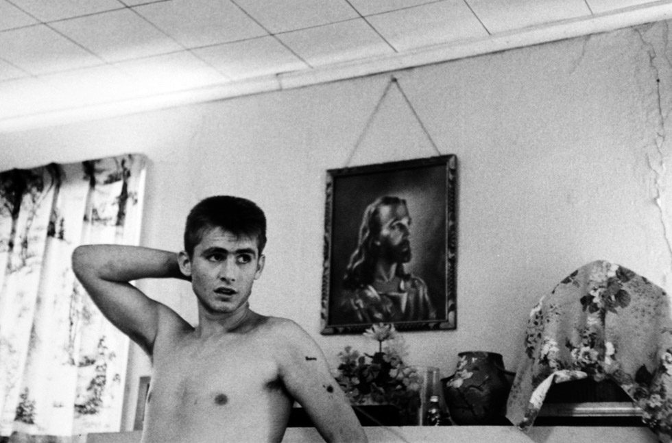 A black-and-white photograph of a shirtless young man in front of a mantle with a painting of Jesus displayed above it.
