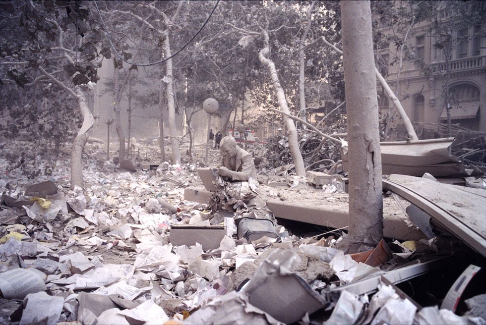 This is a color photograph of a statue pf a businessman on a laptop with the whole scene covered in white dust and sheets of paper and other detritus.