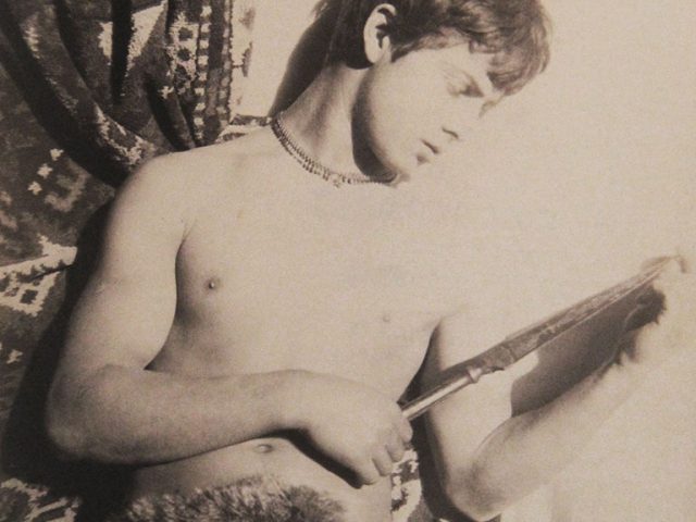 This is a sepia-colored black-and-white photograph of a shirtless boy in a fur loincloth.