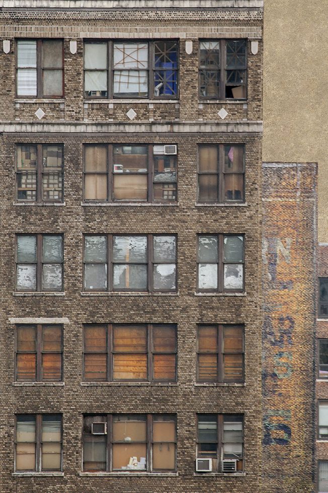 A photograph of the windows in a building, some are boarded up, some taped up, others with peeling paint