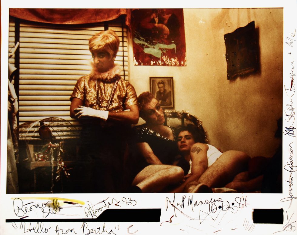 This is a color photograph with written inscriptions in the margins picturing three men in various states of drag in a small interior space.