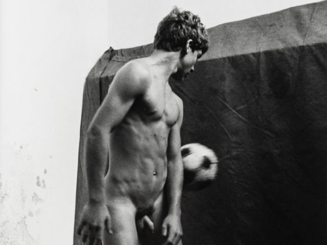 This is a black-and-white photograph of a nude male playing with a soccer ball.