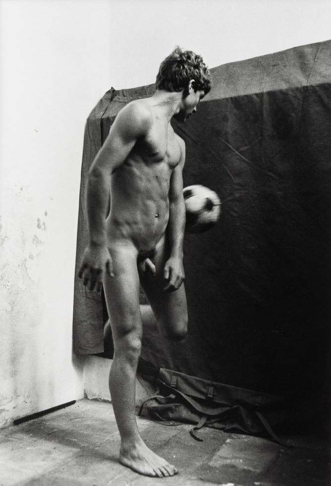 This is a black-and-white photograph of a nude male playing with a soccer ball.