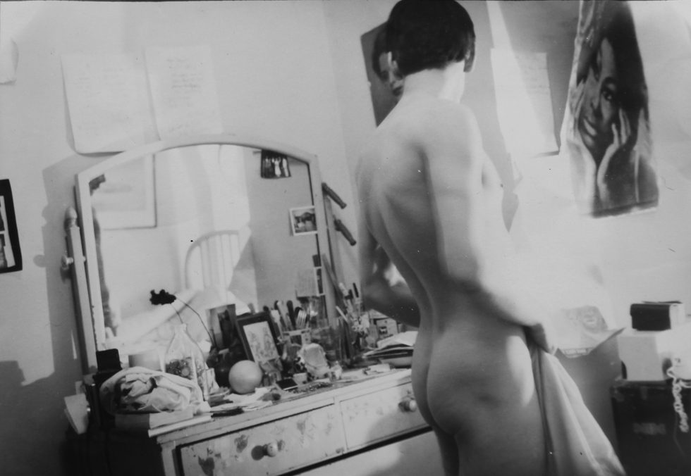 This is a black-and-white photograph of a naked young man in a bedroom in front of a dresser with a mirror.