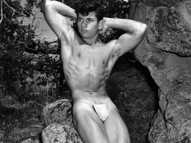 A black-and-white photograph of a bodybuilder in white trunks posing against rocks.