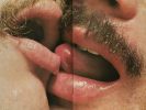 This is a photograph with a seam down the center like a centerfold in a magazine picturing in close up two men with moustaches French kissing.